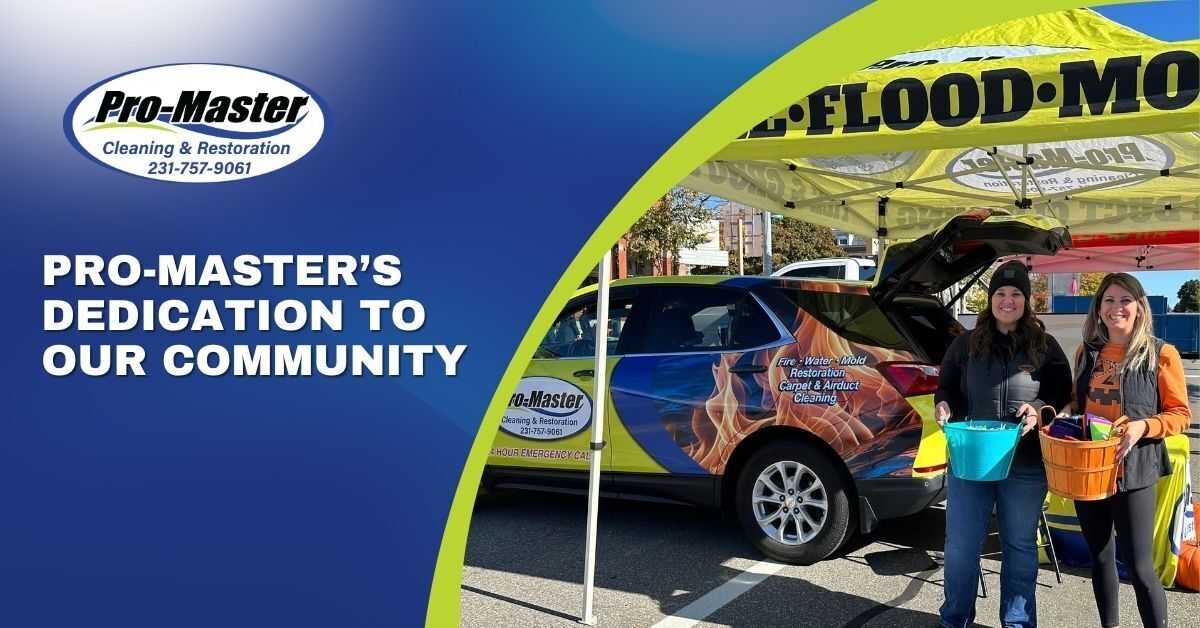 Promotional image for Pro-Master Cleaning & Restoration featuring two women at an outdoor community event. They are smiling and holding buckets filled with small, wrapped items. Behind them is a branded Pro-Master vehicle with graphics and text promoting services like fire, water, and mold restoration, as well as carpet and air duct cleaning. A yellow tent with reversed text and a small orange pumpkin on the ground suggest a fall or Halloween-themed event. The scene highlights Pro-Master's community engagement and dedication.