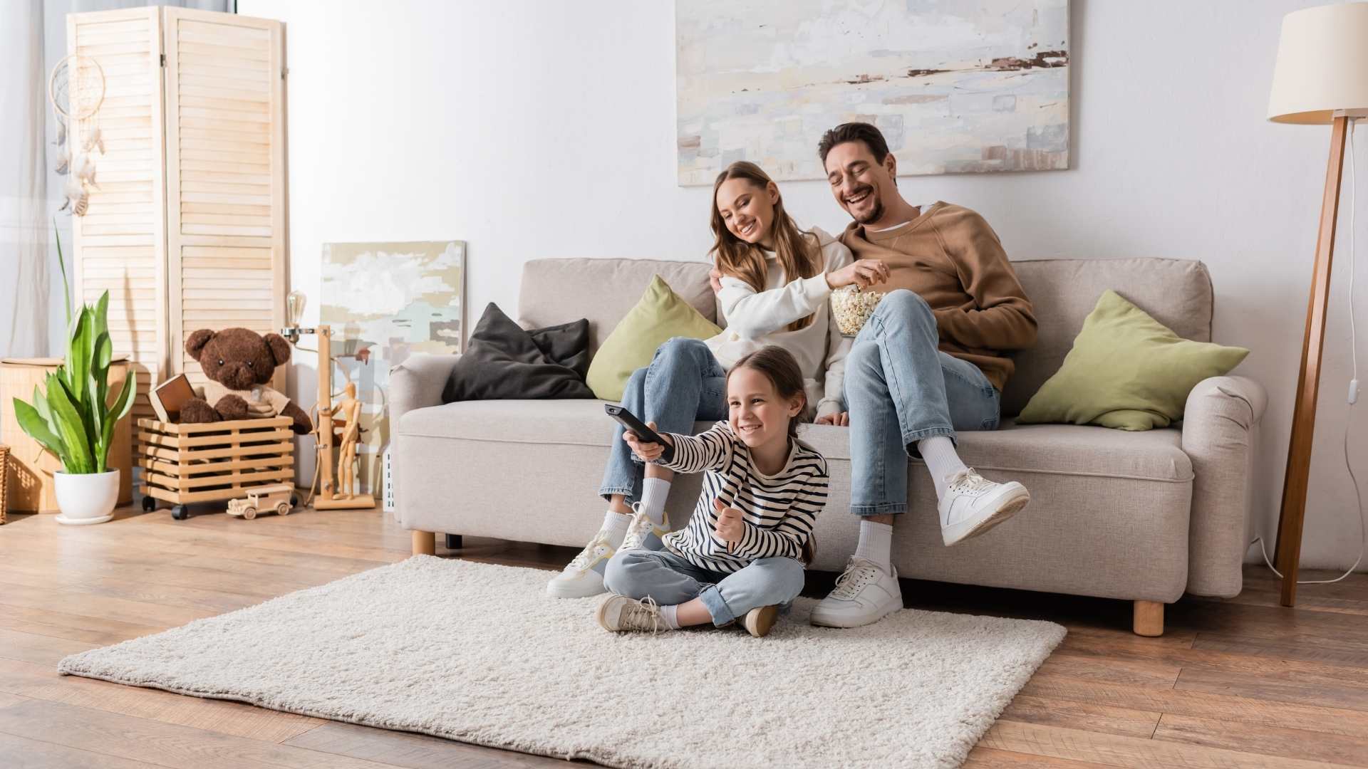 A happy family of three, mother, father, and daughter, relaxing in a modern, brightly lit living room. The daughter sits cross-legged on a shaggy rug, holding a TV remote and smiling, while the parents sit on a light-colored sofa, sharing a bowl of popcorn. The room features wooden flooring, abstract wall art, and cozy decor, creating a warm and inviting atmosphere.